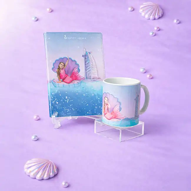 Mermaid-themed mug and notebook on a purple background with shell decorations