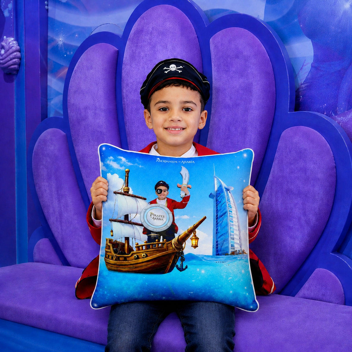 A young boy wearing a pirate hat sits on a purple shell-shaped couch, holding a custom printed blue pillow featuring a pirate ship, a seagull, and the Burj Al Arab hotel logo.