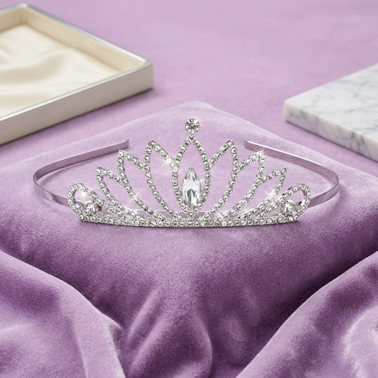 A silver tiara headband adorned with clear rhinestones and a central marquise crystal, displayed on a purple velvet cushion with a jewelry box in the background.