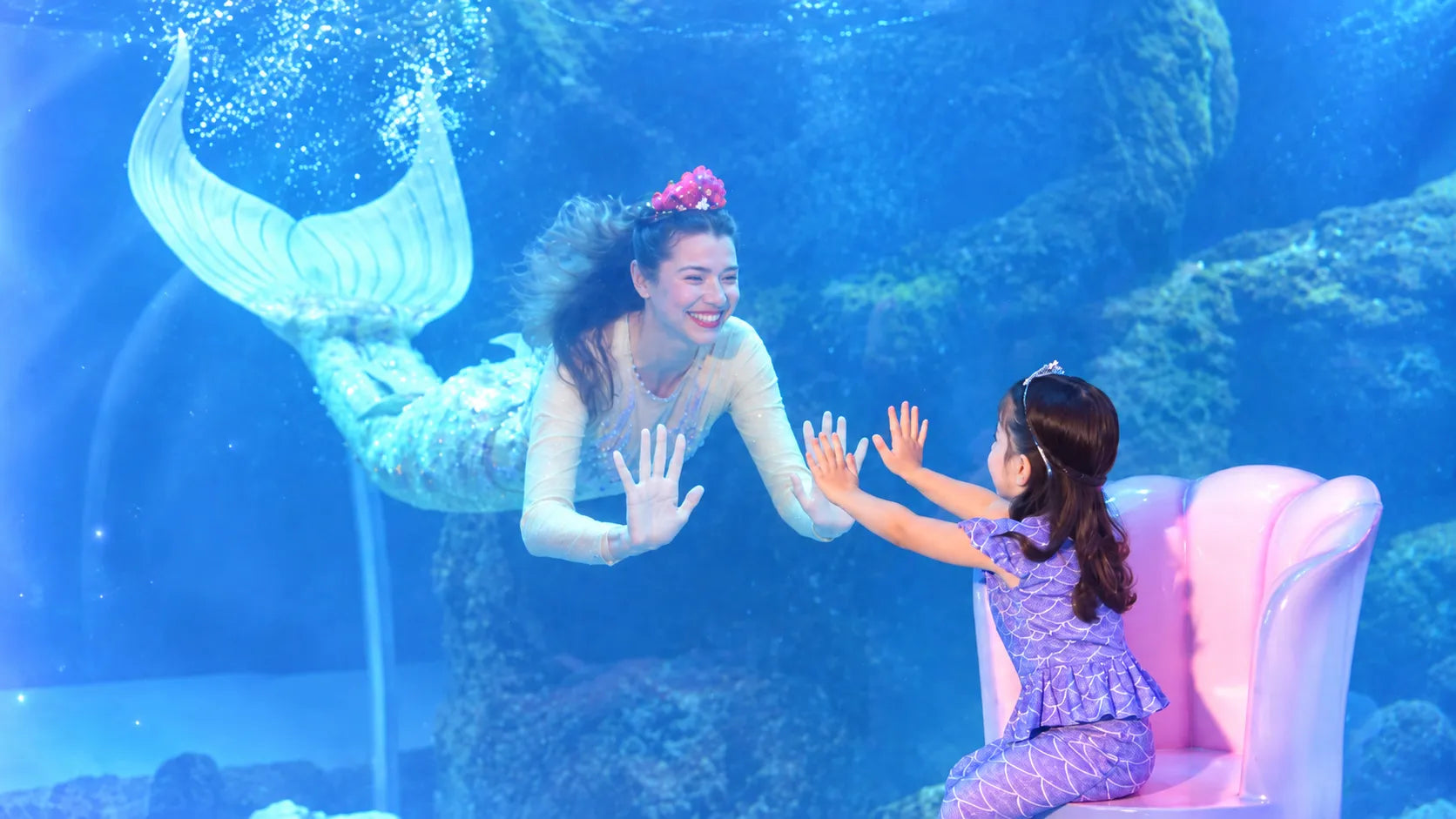 A mermaid and a little girl touching hands through the aquarium glass