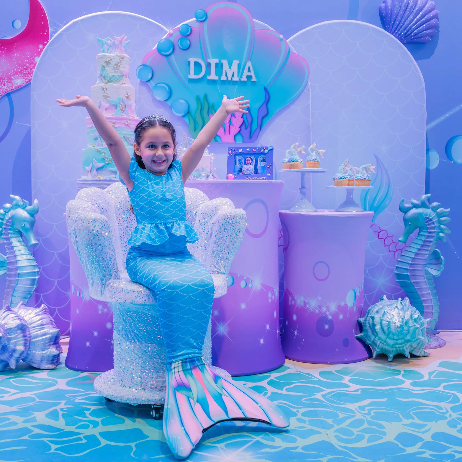 Child in mermaid costume sitting on a chair with decorative ocean-themed backdrop and 'DIMA' text.