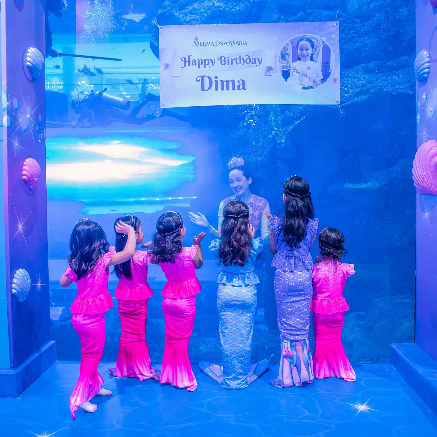 Children in mermaid costumes standing in front of a blue screen with a 'Happy Birthday Dima' sign.