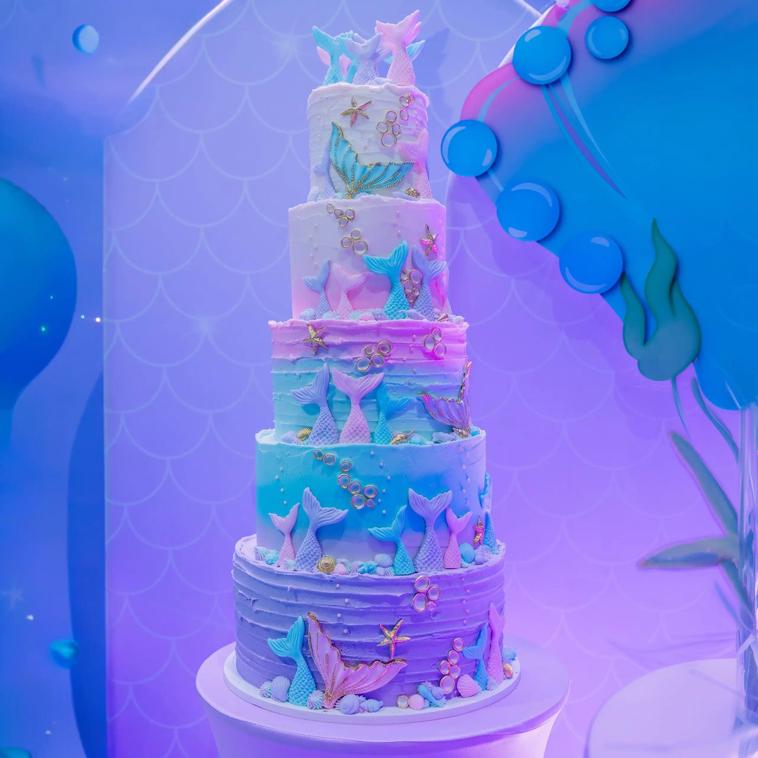 Multilevel cake with mermaid-themed decorations against a blue and purple background