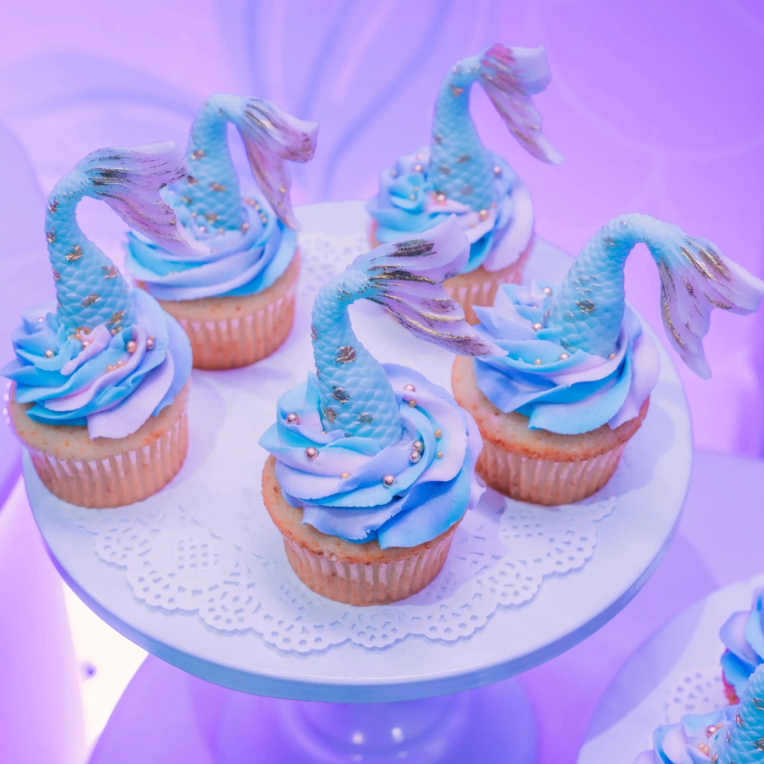 Cupcakes with mermaid-themed decorations on a white cake stand against a purple background