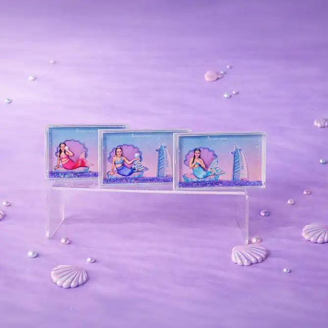 Three mermaid-themed cards on a stand with seashells and pearls on a purple background