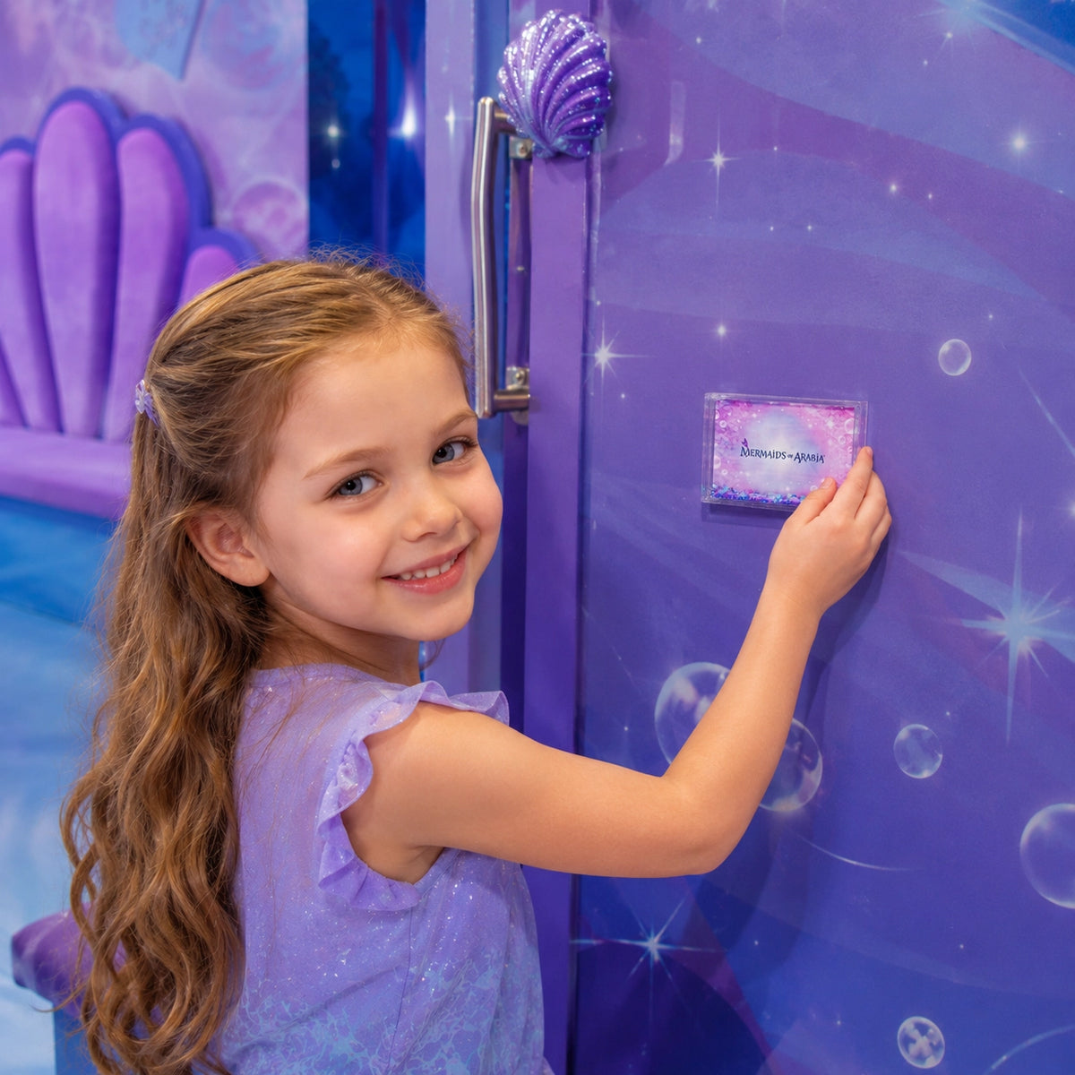 A young girl with long light brown hair smiles while touching a small rectangular purple device mounted on a matching purple door. The door has a shell handle and sparkly star graphics, and the girl is wearing a lavender dress.