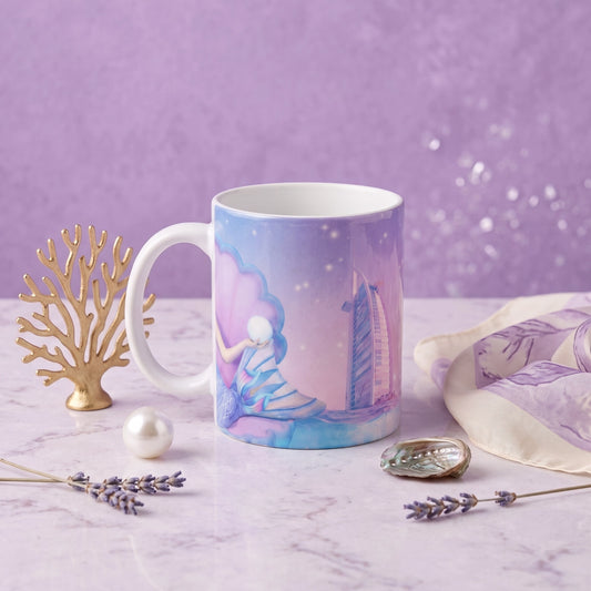A ceramic mug with a pastel mermaid and sailboat illustration, styled on a marble surface with lavender sprigs, a gold coral ornament, a pearl, and a matching purple fabric against a purple background.