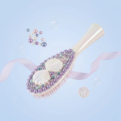 A decorative hairbrush with a pale pink handle and bristles, embellished with white shell motifs, lavender pearls, iridescent beads, and small rhinestones, floating on a light blue background with ribbon and bubble props.