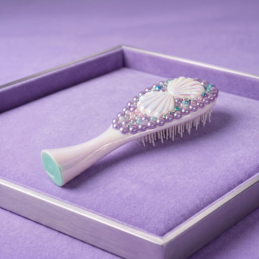 A decorative hairbrush with a pale pink handle, white shell-shaped embellishments, and iridescent pearl-like beads, displayed inside a lavender-lined presentation box.