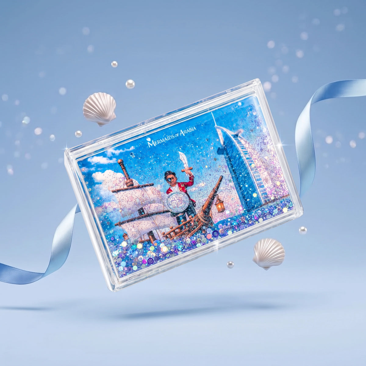 A clear rectangular glitter box featuring a printed scene with a ship, a person in a red outfit, and the text 'Mermaid of Atlantis,' floating on a light blue background with shell props and a ribbon.