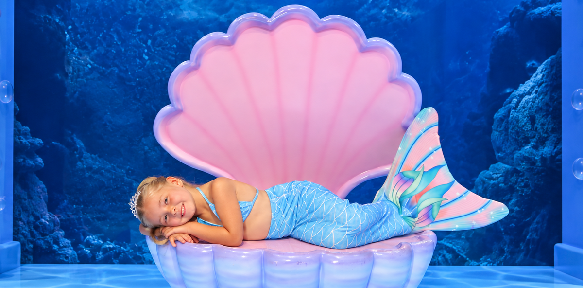 A young girl lies inside a large pink and blue clam shell prop on a themed underwater set with blue coral backdrop, purple columns, and shell decorations.