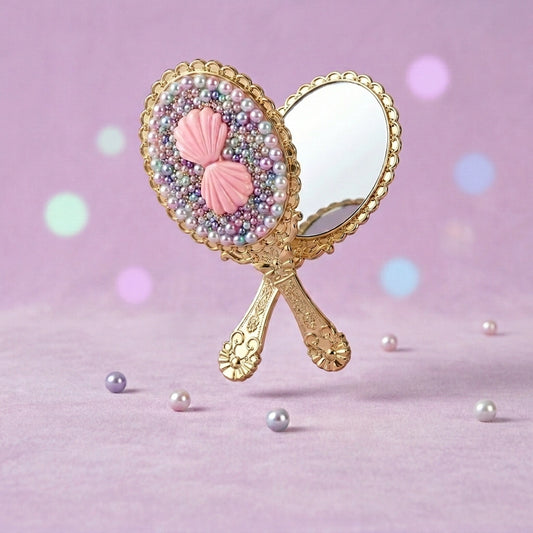 A decorative handheld mirror with an ornate gold handle and a round mirror face embellished with pastel pearls and pink seashell motifs, set against a soft lavender background with scattered faux pearls.