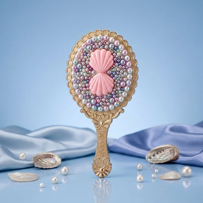 An ornate handheld mirror with a gold-toned handle and scalloped frame, decorated with multicolored faux pearls and a pink shell motif in the center. The mirror is styled on a light blue surface with draped blue satin fabric and scattered real pearls and shell fragments.