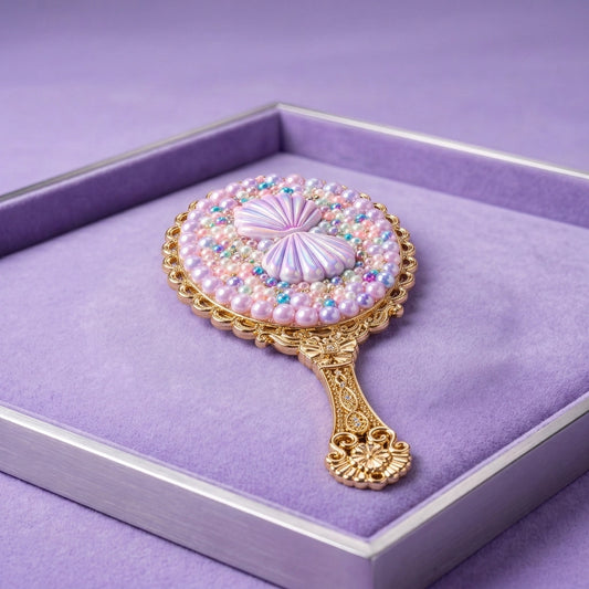 An ornate round hand mirror with a gold filigree handle and a face embellished with pink pearls, iridescent beads, and a central lavender satin bow, displayed inside a lavender velvet-lined presentation box.