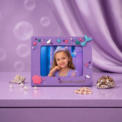 A purple rectangular picture frame with mermaid-themed decorations, including a blue tail, starfish, shells, and pearls, displayed on a lavender surface with seashells and a purple draped backdrop. The frame contains a child's portrait.