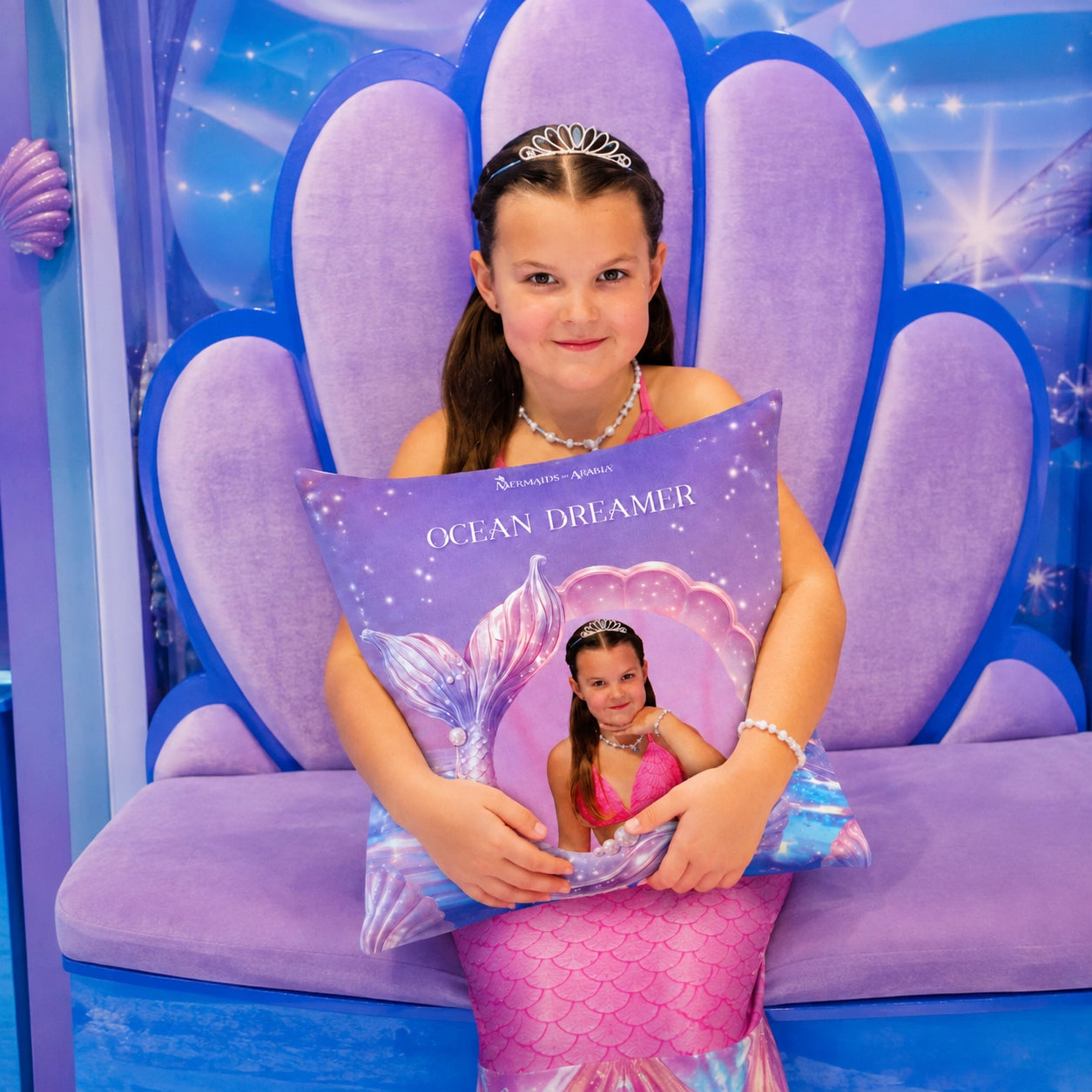 A young girl with a tiara sits on a purple shell-shaped bench holding a decorative pillow printed with the Disney Channel logo and the words 'Ocean Dreamer' with a mermaid tail illustration.