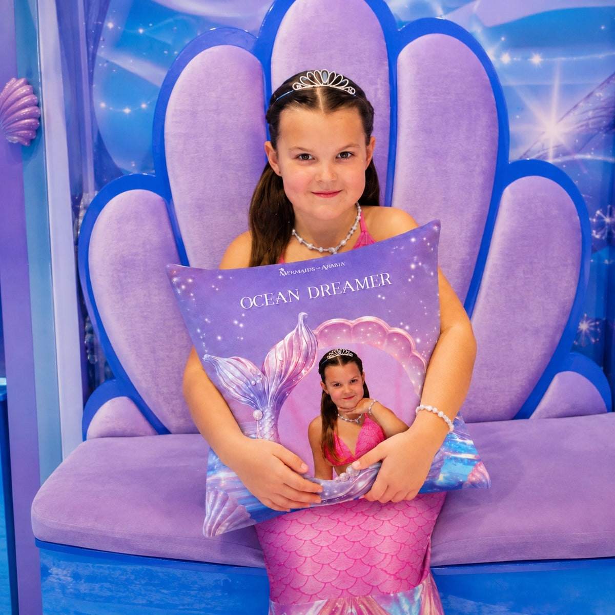 A young girl with a tiara sits on a purple shell-shaped bench holding a decorative pillow printed with the Disney Channel logo and the words 'Ocean Dreamer' with a mermaid tail illustration.