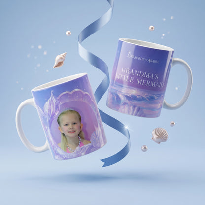 Two custom ceramic mugs with mermaid-themed purple and blue designs, floating against a light blue background with decorative seashells and a blue ribbon. One mug shows a printed portrait of a young girl, and the other has the text 'GRANDMA'S LITTLE MERMAID' and 'MERMAIDS - ARVIA'.
