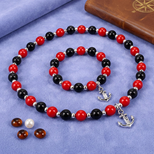 A beaded necklace and bracelet set with alternating red and black round beads, accented by silver-toned metal links and anchor charms, displayed on a purple textured surface with a brown book in the background. A small pearl and two brown beads are placed in the lower left.