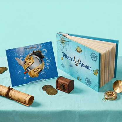 A children's pirate-themed book set displayed on a light aqua surface. The set includes a hardcover book titled 'Pirates of Arabia' with a blue cover, a smaller blue card reading 'I am Brave,' a rolled parchment scroll, gold coins, a small wooden treasure chest, and a compass.