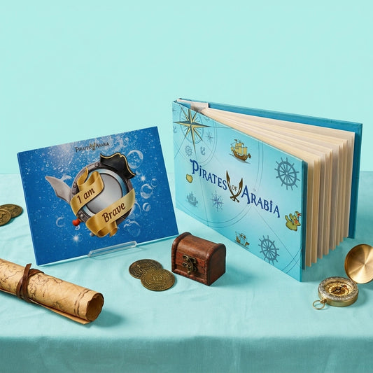 A children's pirate-themed book set displayed on a light aqua surface. The set includes a hardcover book titled 'Pirates of Arabia' with a blue cover, a smaller blue card reading 'I am Brave,' a rolled parchment scroll, gold coins, a small wooden treasure chest, and a compass.