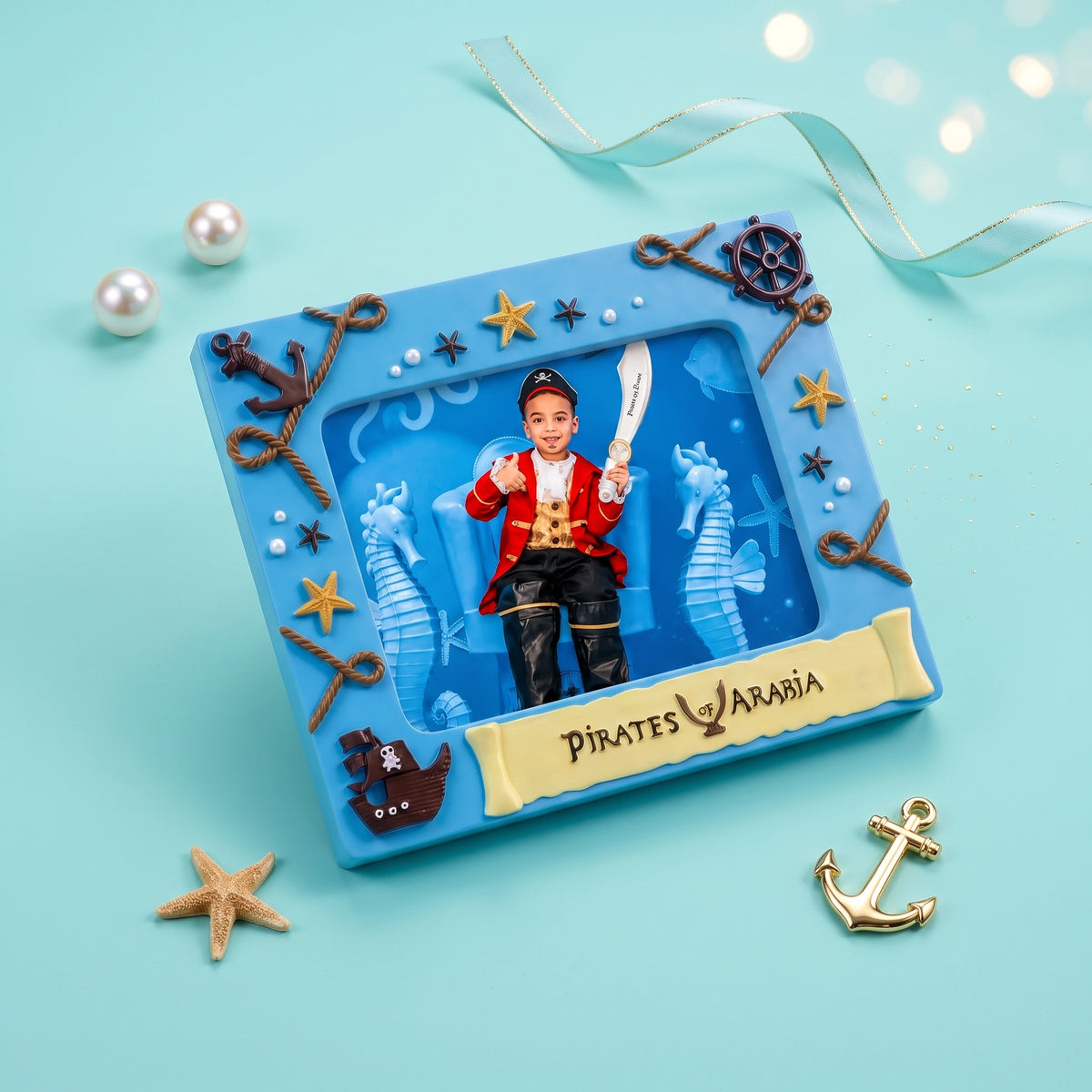 A square blue pirate-themed keepsake box or plaque with a photo window showing a child in a red and black pirate outfit holding a sword. The box is decorated with rope-style anchors, starfish, seahorse cutouts, and a gold banner reading “PIRATES AARON.