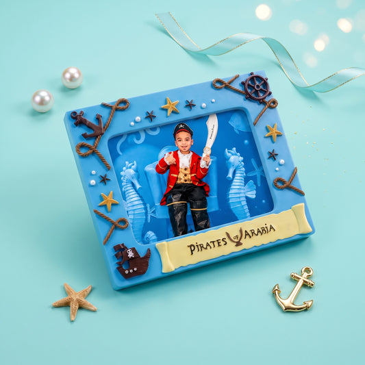 A square blue pirate-themed keepsake box or plaque with a photo window showing a child in a red and black pirate outfit holding a sword. The box is decorated with rope-style anchors, starfish, seahorse cutouts, and a gold banner reading “PIRATES AARON.
