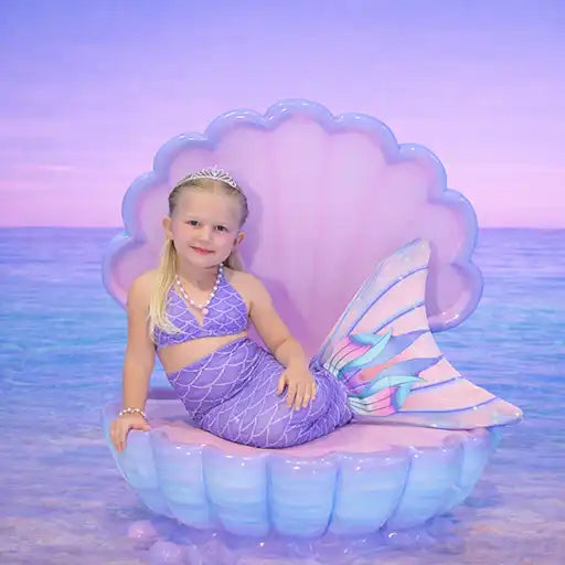Child in a mermaid costume sitting on an inflatable shell with a pastel ocean background