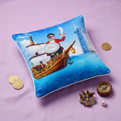 A square decorative throw pillow with a colorful printed design of a pirate ship and a person on deck, styled on a lavender surface with small nautical props including coins, a compass, an anchor, and a pearl.