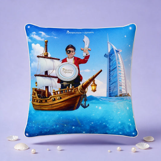 A square decorative throw pillow featuring a colorful pirate-themed illustration with a ship, a person in a red coat holding a sword, and the Burj Al Arab hotel on a blue ocean background. The pillow is photographed against a lavender studio backdrop with small seashell props around it.