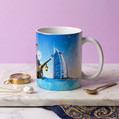 A ceramic mug featuring a printed illustration of a man with a telescope and the Burj Al Arab hotel in Dubai, set on a marble tray with a gold pocket watch, pearl, and spoon against a lavender background.