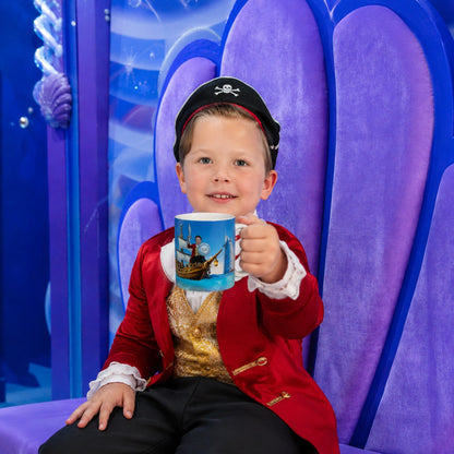 A young boy dressed as a pirate sits on a purple throne-like chair, holding up a blue printed mug with a pirate ship illustration. The background is a bright blue and purple themed set.