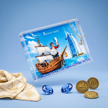 A clear acrylic keepsake box with a printed Dubai-themed illustration, shown on a light blue background with a gold anchor pendant, blue gemstones, gold coins, and a cream scarf with gold embroidery.