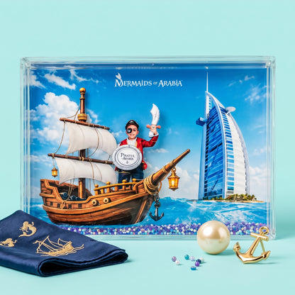 A framed art print titled 'Mermaids of Arabia' displayed on a light blue background, featuring a pirate ship, a mermaid character, and the Burj Al Arab hotel. A navy blue fabric with gold embroidery, a pearl, and gold anchor accents are arranged in front of the frame.