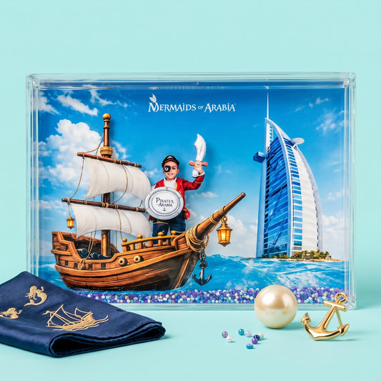 A framed art print titled 'Mermaids of Arabia' displayed on a light blue background, featuring a pirate ship, a mermaid character, and the Burj Al Arab hotel. A navy blue fabric with gold embroidery, a pearl, and gold anchor accents are arranged in front of the frame.
