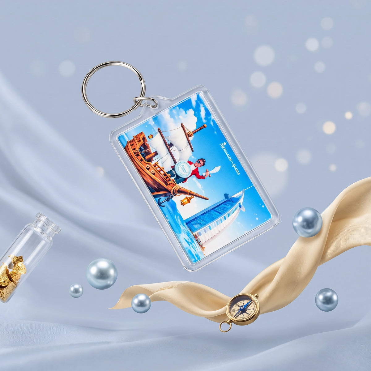 A clear acrylic keychain with a silver metal split ring, featuring a colorful printed illustration of a pirate ship and a sailor on a blue background. The keychain is shown floating against a soft light blue backdrop with decorative pearl-like spheres, a small glass vial with gold coins, and a beige ribbon with a compass.