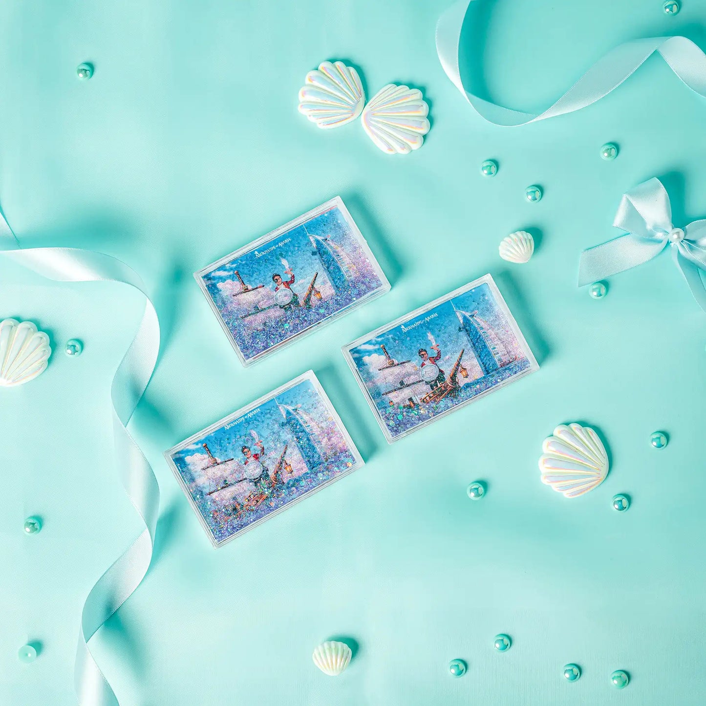 Three decorative cards with mermaid designs on a turquoise background with shells and ribbons.