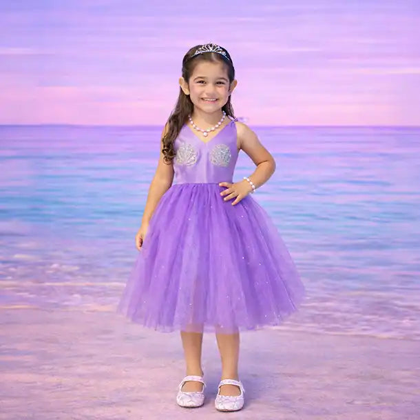 Young girl in a purple dress with silver accents standing on a beach with a pastel sky.