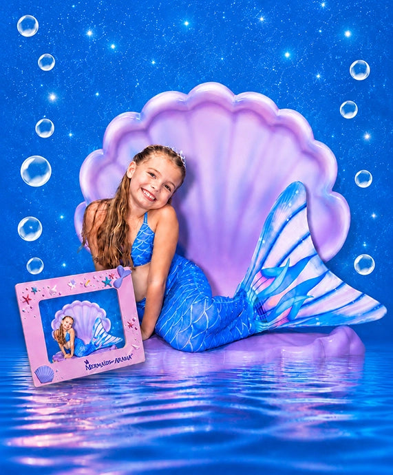 A young girl with long light brown hair sits in a stylized underwater scene wearing a blue mermaid tail and matching blue top, posed in front of a large purple shell prop. A pink framed picture with a mermaid illustration rests beside her.