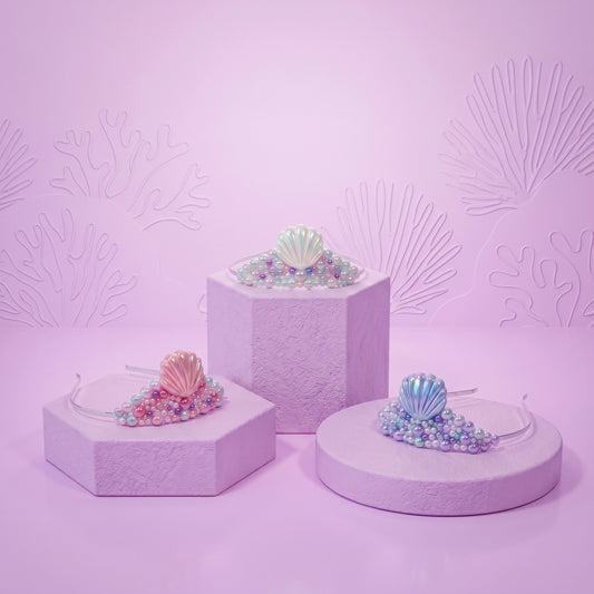 Three clear plastic headbands decorated with iridescent shell and bead designs, displayed on pastel lavender pedestals against a matching lavender background with embossed coral patterns.