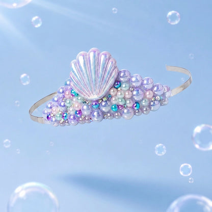 A mermaid-style headband with a large iridescent shell motif and clusters of pearl-like beads in lavender, white, and blue, shown against a light blue background with floating bubbles.