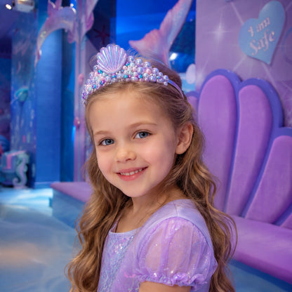 A young girl with long wavy light brown hair smiles while wearing a purple mermaid-themed costume dress and a matching purple seashell tiara in a colorful indoor play area.